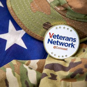 An american flag behind a camo patterned hat and shirt. A badge with "Veterans Network Comcast" on top.