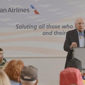 person stands at a podium in front of a crowd. American Airlines logo behind them