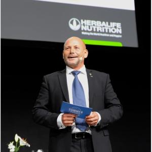A person standing on a stage, holding a blue card. "herbalife nutrition" logo behind them.