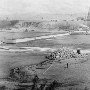 old aerial photo of a river with a dam across it. a building on the distant hill.