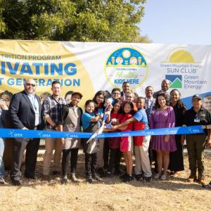 Group of people, including children, standing behind a ribbon, in front of a shipping container farm.