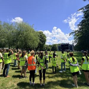 College program participants wearing high-visibility safety vests stand in a grassy field while receiving a festival operations briefing with an outdoor stage visible in the distance.