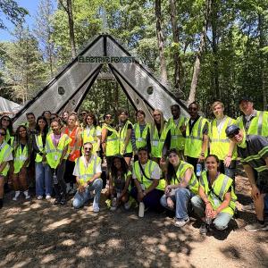 A group of college program participants wearing high-visibility safety vests poses for a photo beneath the Electric Forest entrance structure in a wooded festival setting surrounded by trees.