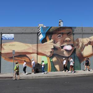 Volunteers walk past a large colorful mural while participating in a neighborhood cleanup and beautification effort on a city street.