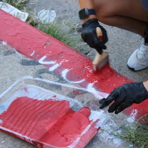 Close-up of volunteers wearing gloves repainting a red curb on a city sidewalk using brushes and a paint tray filled with red paint.