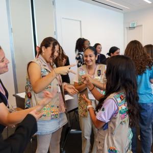 Participants stand in a circle indoors, engaging in an interactive group activity during a workshop, with a presentation screen visible in the background.