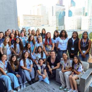A large group of participants gathers on an outdoor terrace with city buildings in the background, posing together during a group activity.