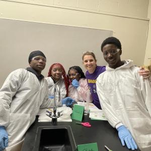 A woman in a purple Curiosity Labs™ MilliporeSigma t-shirt stands with students in lab equipment as they conduct experiments. 
