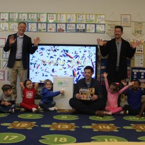 Two men stand and wave smiling, while children sit around an oversized prop light switch cheering