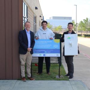 Three men standing around blue sign smiling, with one of them holding an oversized prop light switch
