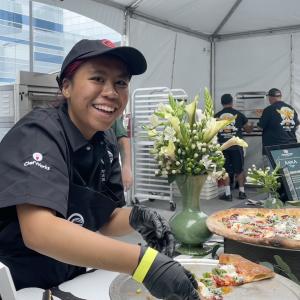 Food service worker wearing gloves prepares pizza slices at a festival booth, with a decorated pizza display on the counter.