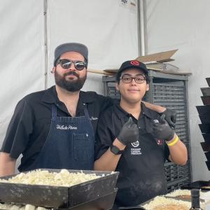 Two food service workers stand behind a pizza prep station with trays of shredded cheese inside a tent.