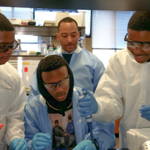 Dr. Arik King in a lab working with three young men of color. All are wearing lab coats and protective eye ware. 