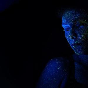 a dark space, a face lit by blue-light on the right, green paint spatters on the face