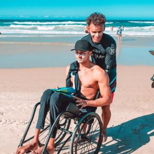 A person in a wet suit helping another in a wheelchair on a beach.