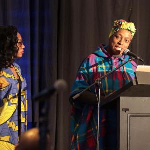 Bridget Ndagaano and Yvette Raphael on stage. Yvette behind a podium with microphone.