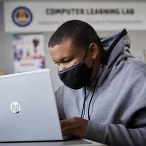 a student in a mask and gray hoodie leans close to an HP laptop screen