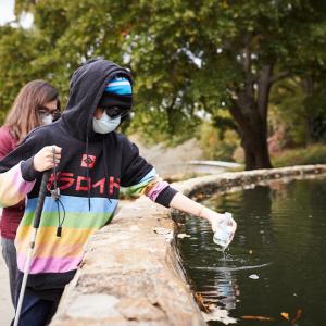 a student wearing a mask and dark sunglasses dips a water bottle into a pool