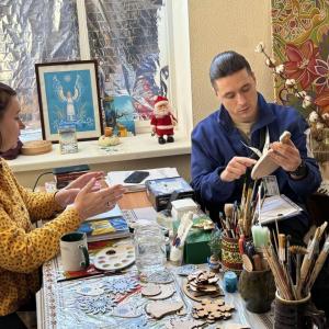 A woman in a yellow shirt gives a lesson to a man in a blue jacket. The are surrounded by painting materials.