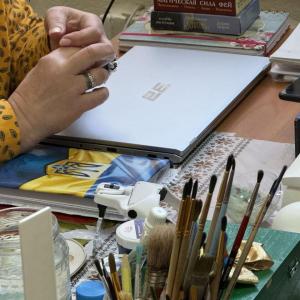 A woman's interlaced fingers hover over a closed laptop. Paintbrushes are on the desk.