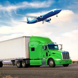 A green semi truck on the road and a jet plane in the sky above it.