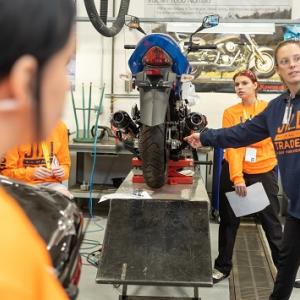An adult pointing to a spot on a raised motorcycle as students look on.
