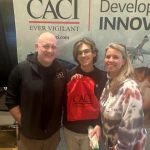 A group of three pose in front of a CACI banner, holding a CACI bag.