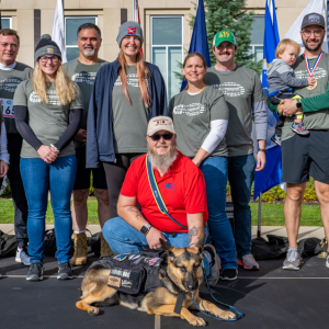 A group of participants, a person kneeling in front of them with a service dog. Flags in the background.