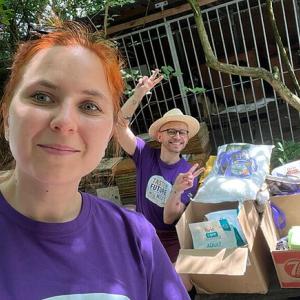 Karol in the background of a selfie with another teammate. Packed boxes of supplies on the ground next to them.