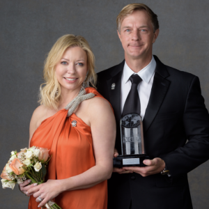 Two people stood next to each other smiling, one person holding flowers and the other holding an award