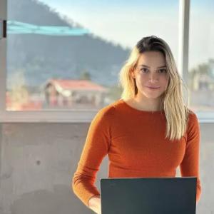 Karla Huerta with an open laptop sitting on a bench in front of a large window.