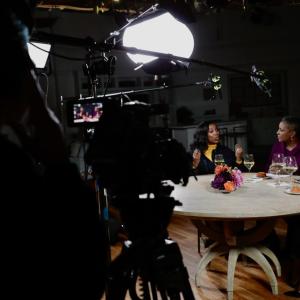 QVC Hosts Vanessa Herring, Jayne Brown, Monifa Days and Stacey Rusch are seated at a table with a television camera filming them while on-set at QVC Studios.