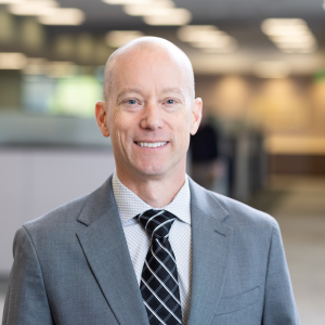 Man wearing a grey suit, white checkered shirt, and black-and-white striped tie, standing in a modern office with cubicles and ceiling lights.