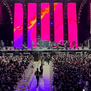 A large performance stage with digital screens, a large seated audience, and ushers in the aisle.