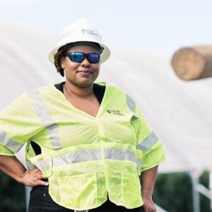 LaQuitta Ghent next to large equipment, wearing high-vis vest at hard hat. One hand on a hip.