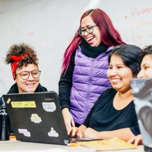 Three smiling people seated, looking at a laptop, a fourth behind them. A whiteboard with writing on it in the background.
