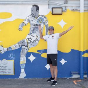 LA Galaxy player Landon Donovan poses with his arms outreached in front of a yellow, blue and white mural of him in a LA Galaxy jersey kicking a soccer ball. 