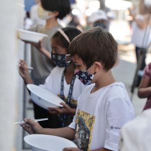 Elementary students wearing masks, each hold bowls of paint and brushes as they stand in a line and finish the LA Galaxy mural at their school. 