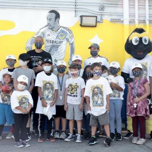Students in Landon Donovan t-shirts and masks crowd around LA Galaxy player Landon Donovan in front of a yellow, white and blue mural of Landon Donovan in an LA Galaxy jersey. 