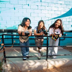 Three young people hold cameras and stand together along a railing in front of a bright blue brick wall.