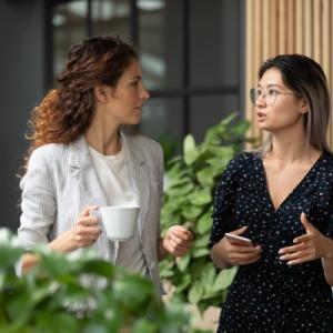 Two people talking, one with a white cup in their hand, the other has a phone. In an office setting with house plants in front and behind.