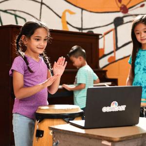 children standing in front of a laptop, one behind them playing on a piano. Wall behind them with a painted mural of floating music notation.