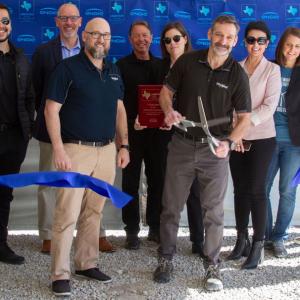 A group of people pose as one uses a large scissors to cut a blue ribbon.