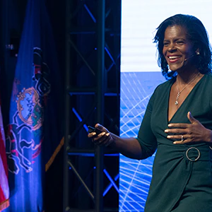 Lynnette speaking with hand gestures, two flags to the left and a large digital screen behind.
