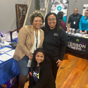Three people pose for the camera in front of tables set up for different organizations.