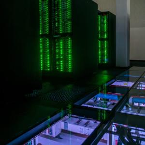 A dark upper room with green-lit large electronics. A clear section of floor showing a room below.