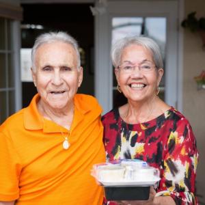 two elderly people on a front porch, one holding a packaged meal