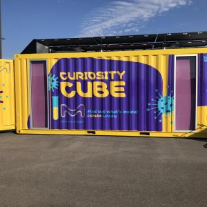 A yellow and purple shipping container with the words "Curiosity Cube" on the side showcase an environment dedicated to expanding science education