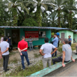 A group of people standing outside a small building surrounded by tropical trees. All listening to a speaker.