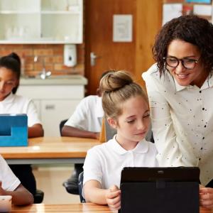 Classroom of students at long desks, each with an electronic device. An adult looks over one of their shoulders
